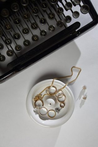 An assortment of gold and silver jewelry adorns a round ceramic dish, featuring El Camino College part-time Faculty Librarian Catherine Bueno's sterling silver earrings, showcased beside an old-fashioned typewriter. (Raphael Richardson | Warrior Life)