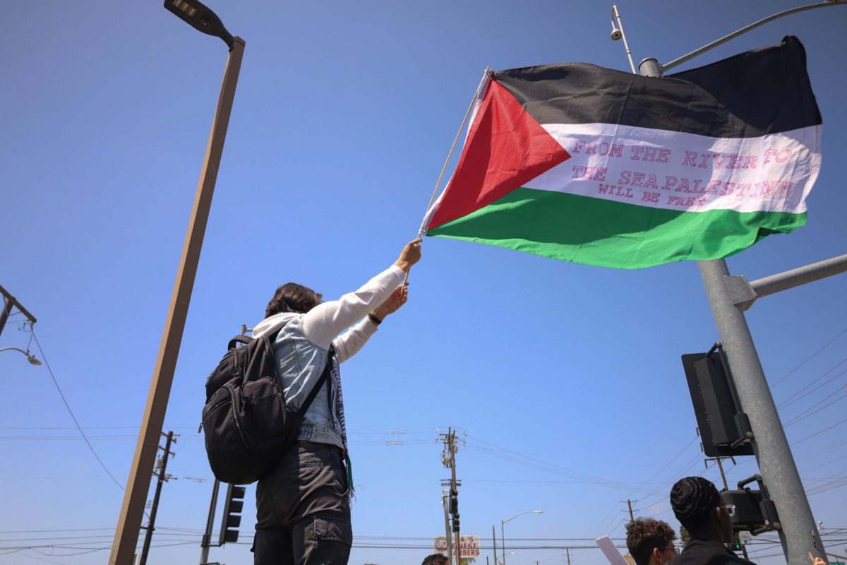 A protester waves a Palestinian flag, with a phrase written on it, on the corner of Manhattan Beach and Crenshaw Boulevards on Tuesday, May 21 as part of a student-led Teach-In for Palestine event that turned into a march. Marching students and employees congregated on the corner outskirts of El Camino as some passing cars honked their horns in support. (Delfino Camacho | The Union)