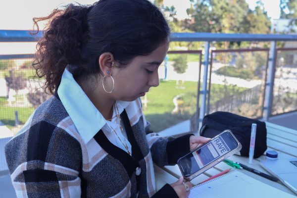 Camila Martinez, 19-year-old biochemistry major, looks at the RAVE alert system page on the El Camino College website through her phone while sitting on a bench outside the Behavioral and Social Sciences Building on Oct. 22. Martinez didn't know about the previous alert system NIXLE and found out about RAVE through emails from the college. She plans on signing up for RAVE text alerts to get information about things happening around campus. (Eddy Cermeno | The Union)