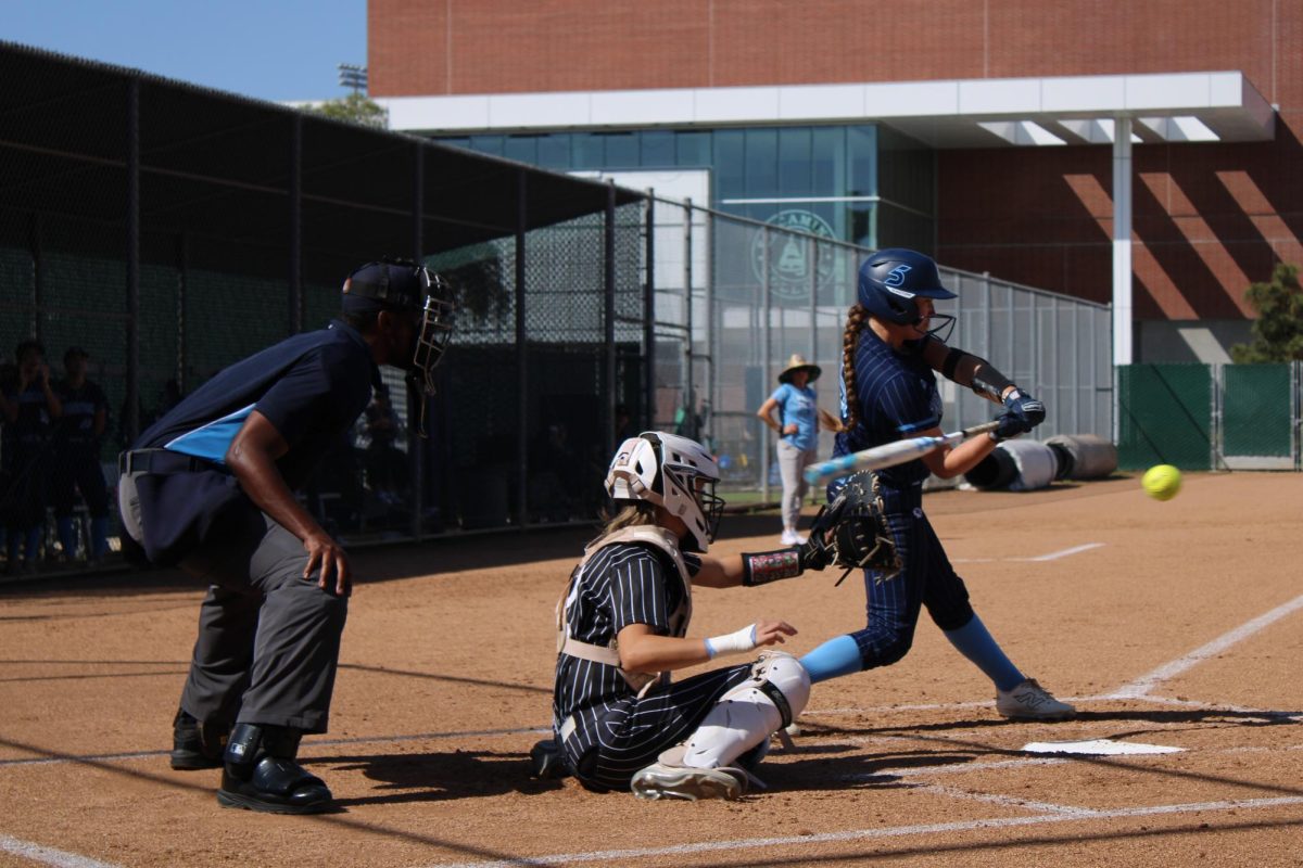 (L-R) Home plate umpire Anthony Banks and Long Beach City College Vikings catcher Zara Mineo watch the ball as El Camino College Warriors second baseman Lilly Lewis takes a swing on the ECC Softball Field Friday, Feb. 28. Lewis gets the single, but is left on base at the end of the first inning. (Elsa Rosales | The Union)